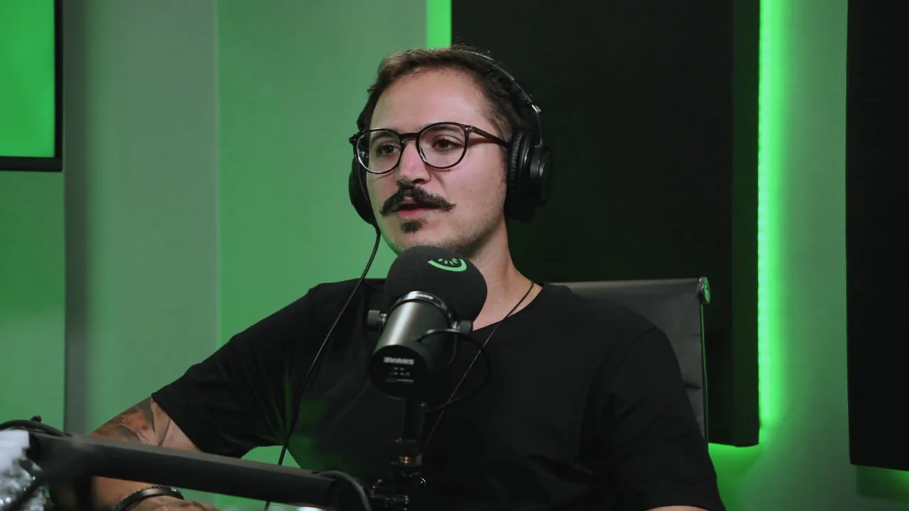Fillipe de fones de ouvido e óculos, falando ao microfone com a marca da Kiwify, no estúdio verde do podcast.